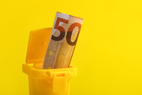 A fifty euro banknote placed in a small yellow trash bin against a vibrant yellow background, symbolizing financial waste or loss.