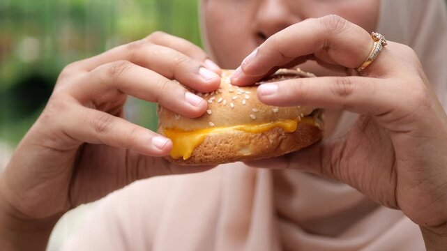 A close up view of a woman in a hijab holding and gently pressing a juicy cheeseburger in her hands outdoors, ready to take a bite