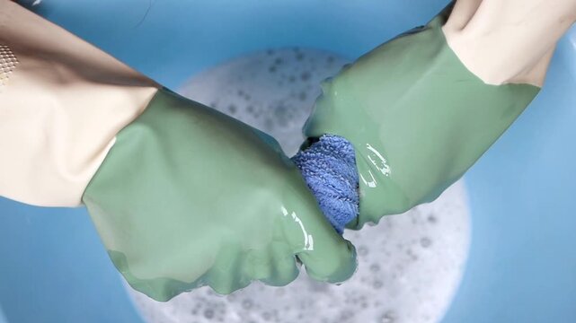 Hands wearing gloves washing blue cloth in soapy water close up