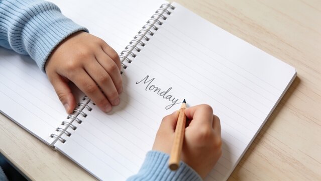 Child writing the word Monday in a notebook with a wooden pencil on a wooden desk