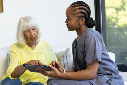 Senior woman in yellow top at home African-American woman aide in gray scrubs sitting with tablet