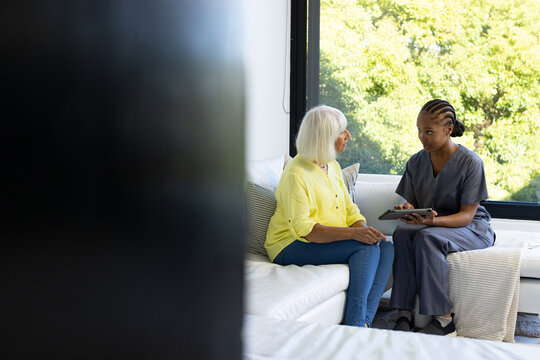 Female senior and African American nurse in gray scrubs sitting on sofa using tablet, copy space