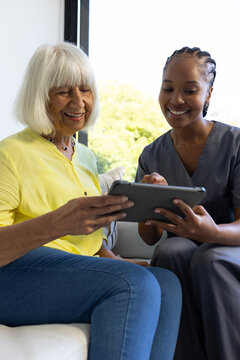 Female caregiver in gray scrubs and senior woman in yellow blouse sitting on sofa pointing tablet