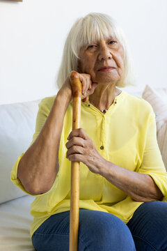 Senior woman sitting on sofa in living room wearing yellow blouse and blue jeans holding cane