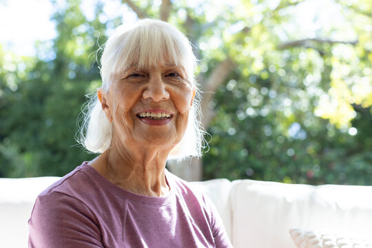 Senior woman sitting on outdoor sofa in backyard, smiling in mauve top with patterned pillow