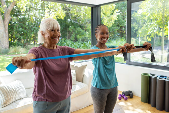 Diverse female friends in workout clothes using resistance bands at home, senior wearing maroon tee
