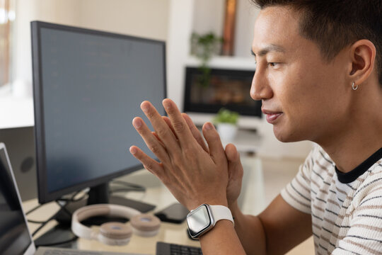 Asian man sitting at home desk wearing striped tee, watch, viewing dual screens, copy space