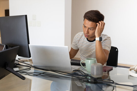 Asian man working on laptop in striped T-shirt with smartwatch at glass desk, monitor, copy space