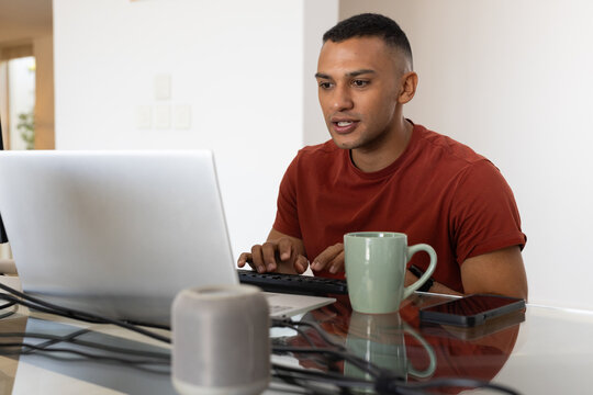 African American man sitting at glass table at home, working on laptop, keyboard, phone, green mug
