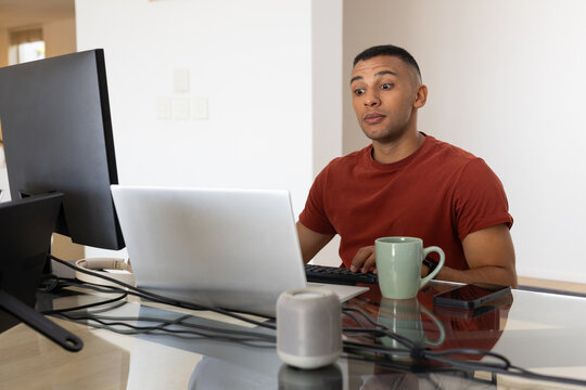 Adult African man sitting at glass desk at home, wearing rust T-shirt and working on laptop