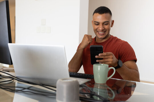 Adult African man sitting at glass desk smiling, checking phone near laptop, green mug, copy space