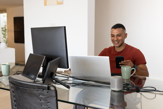 African American man sitting at glass table using laptop, holding smartphone, red tee, copy space