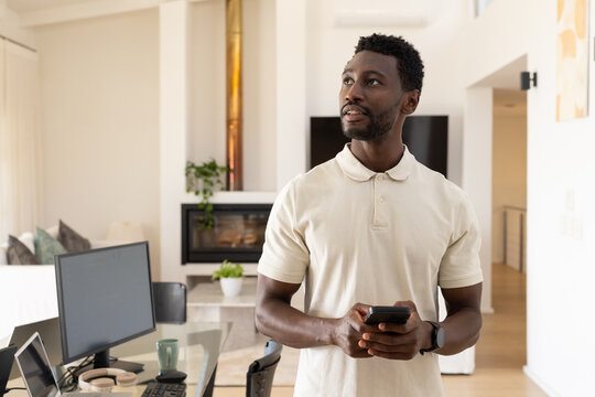 African American man in polo shirt and smartwatch standing in home office holding phone
