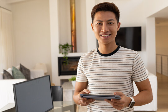 Asian mid-adult man standing in living room home office holding tablet by glass table