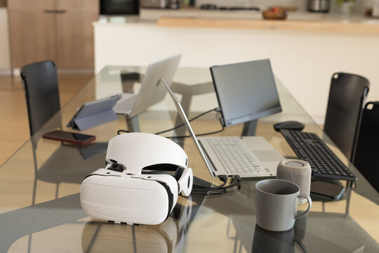 White VR headset is resting on glass table surrounded by laptops, monitor, tablet in kitchen
