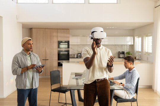 Diverse male coworkers testing white VR headset, using tablet and laptop in open-plan kitchen
