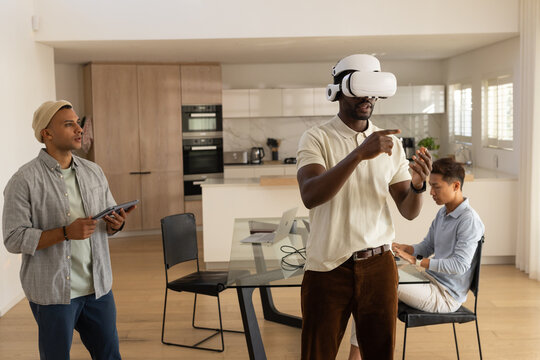 Diverse male friends testing VR headset with controller at glass kitchen table, friend using laptop