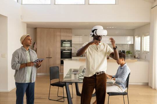 Diverse male coworkers testing VR headset, watching tablet, typing on laptop in kitchen