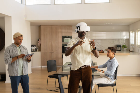 Diverse male coworkers testing white VR headset while using tablet and laptop in kitchen