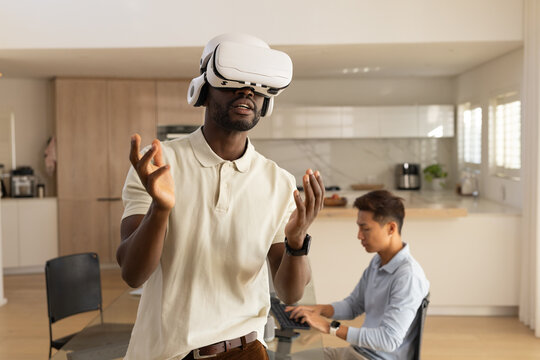 Diverse male friends with white VR headset gesturing while other working on laptop at kitchen table