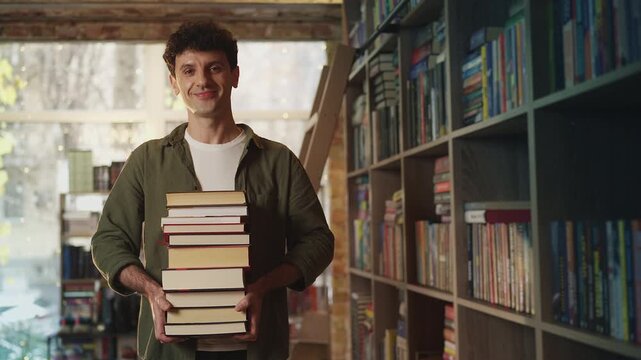 Portrait of happy male librarian holding stacks of books at his workplace