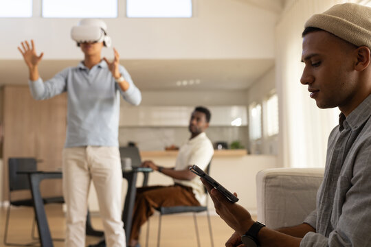 Diverse male friends in open-plan home using phone, wearing VR headset and using laptop, copy space