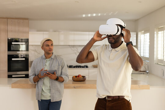 Diverse male friends adjusting white VR headset and holding tablet in kitchen by wooden island