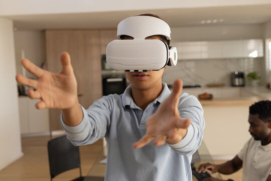 Diverse men sharing space with Asian man wearing VR headset reaching forward at kitchen with laptop