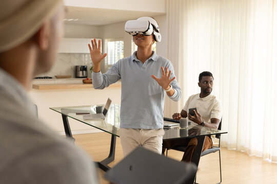 Diverse male team watching Asian man wearing white VR headset at glass table, holding smartphone