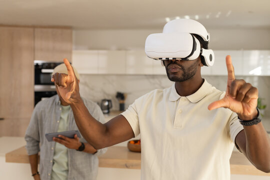 African American men, one wearing white VR headset, gesturing while other holding tablet in kitchen
