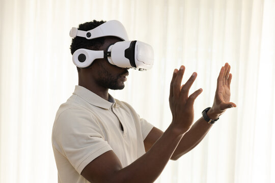 African man wearing beige polo, white VR headset, smartwatch, gesturing hands near sheer curtains
