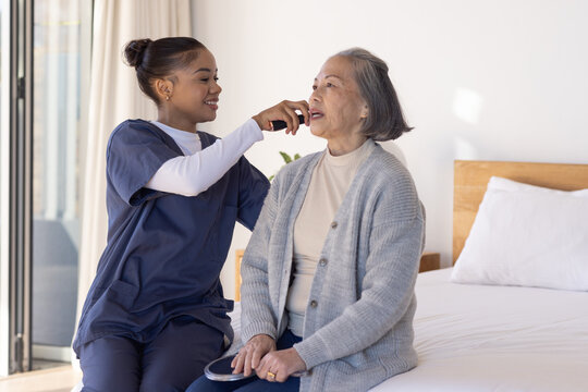 Female Asian senior and African American caregiver in scrubs taking temperature with device at home
