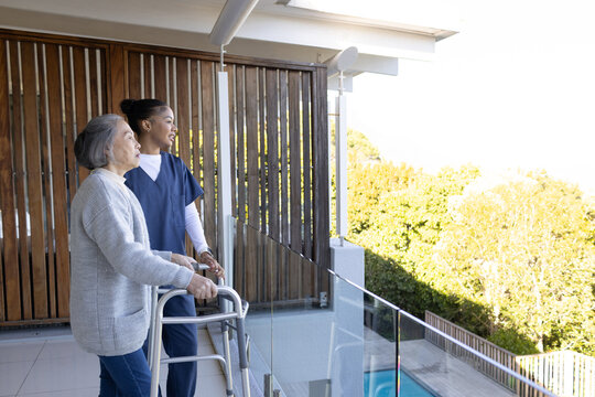 Senior Asian woman using walker, caregiver in scrubs supporting on balcony by glass railing pool