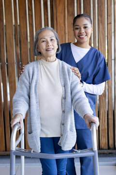 Senior female Asian standing with walker, African American caregiver in scrubs supporting at porch