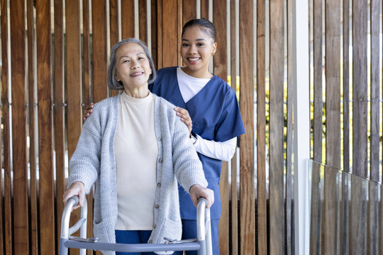 Senior Asian woman using walker on porch while African American caregiver supporting in navy scrubs
