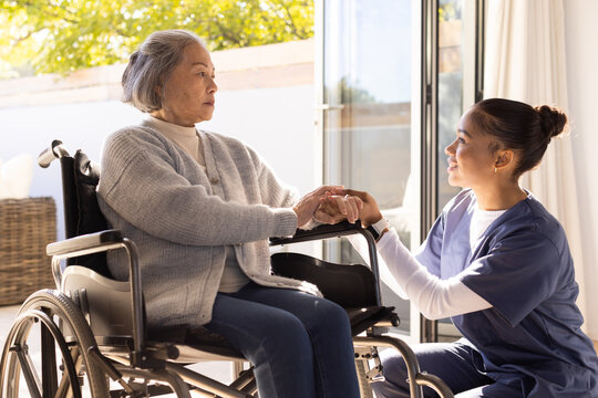 Diverse Senior Asian woman  and  African-American nurse-in-scrubs holding hands by wheelchair at hom
