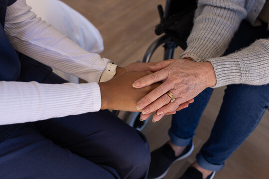 Asian senior woman and African American carer holding hands at home, wheelchair, rings, white watch