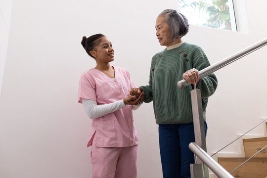 Senior female and Asian caregiver in pink scrubs holding Senior's hand by metal handrail on stairs