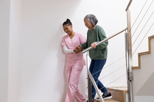 Senior Asian woman descending stairs home, female caregiver in pink scrubs guiding hand on handrail