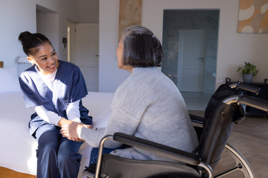 Senior Asian woman sitting in wheelchair holding hands with nurse wearing navy scrubs on bed
