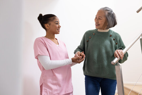 Diverse female caregiver in scrubs holding Asian senior's left hand with ring on stairs, metal rail