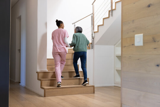 African-American female caregiver in scrubs holding senior Asian woman's hand up wood stairs home
