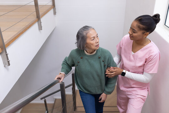 Diverse female nurse in scrubs helping senior in green top climbing stairs at home holding rail