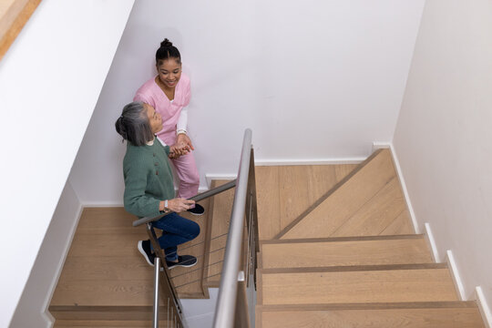 Senior female and female caregiver in pink scrubs descending stairwell, holding metal handrail