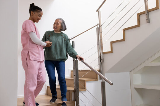 African American caregiver holding Senior Asian woman's hand, guiding down stairs with metal rail