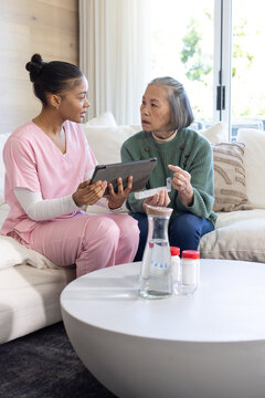 African American caregiver in scrubs sitting on sofa with Asian senior holding tablet and pillbox