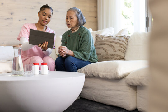 Female caregiver in pink scrubs and Asian senior sitting on sofa holding tablet near pill bottles