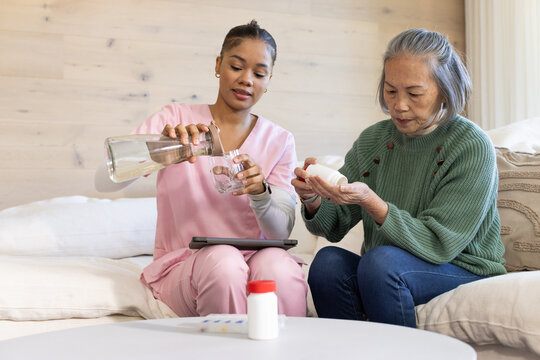 Diverse female carer in scrubs pouring water, tablet on lap, senior woman dispensing pills on sofa