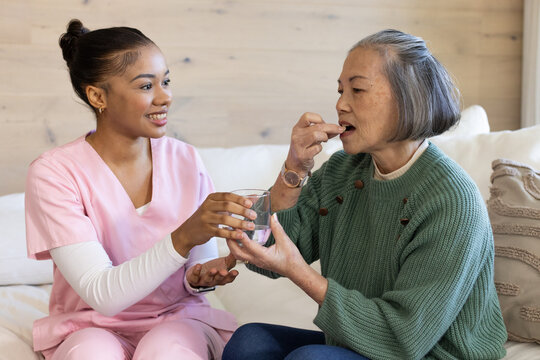 Pink-scrubbed nurse helping senior Asian woman in green sweater taking pill with glass on sofa