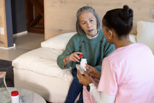 Senior Asian woman, African-American nurse wearing scrubs on sofa holding pill bottle, blister pack